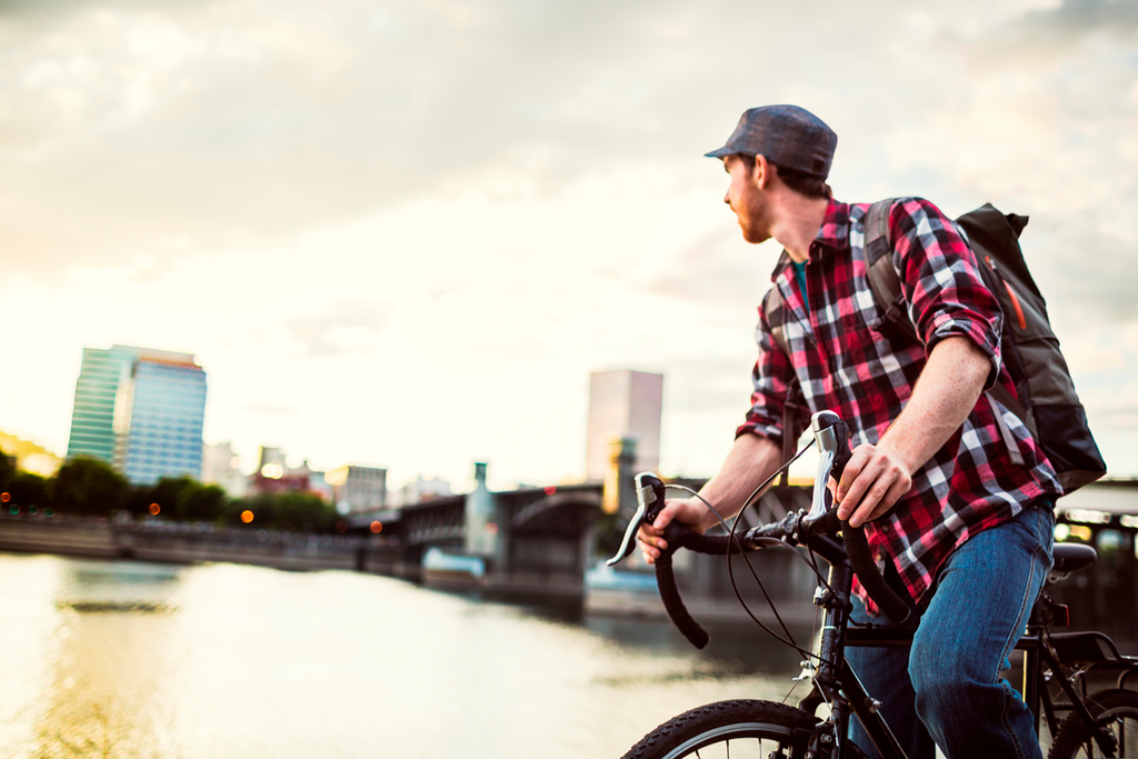 Homme portant un jeans, une chemise et un sac à dos, en vélo dans une ville, longeant une rivière au niveau d'un pont