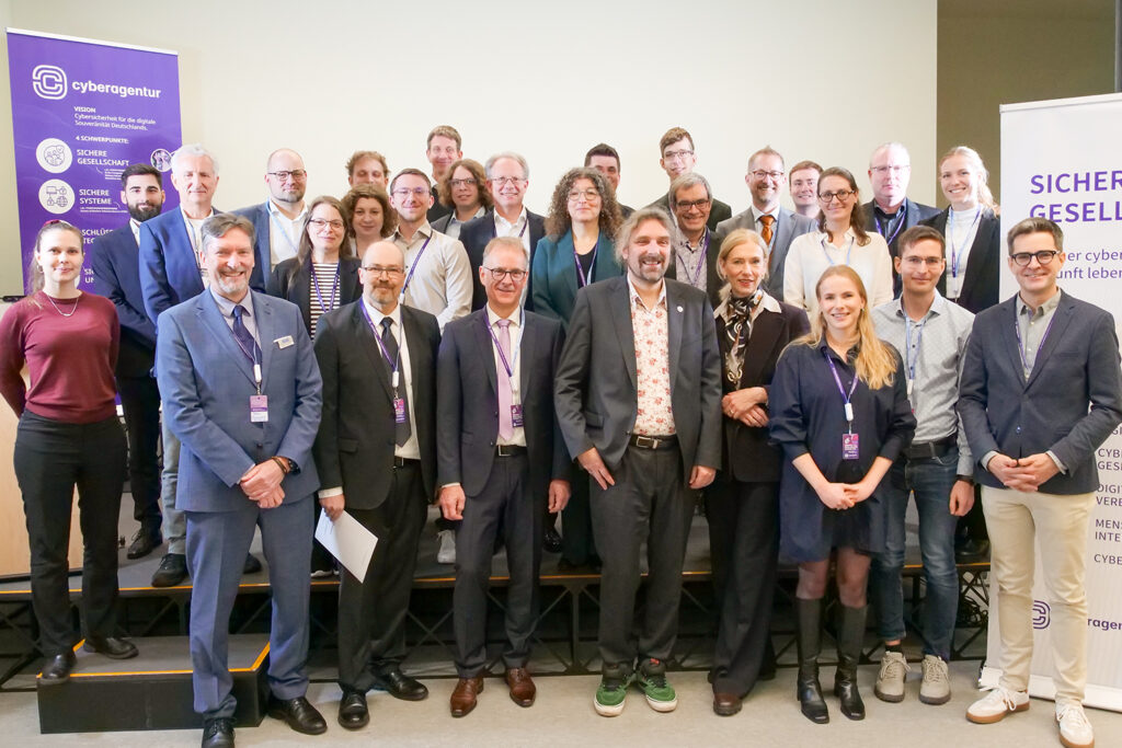 Gruppenfoto mit etwa 30 Personen – Vertreterinnen und Vertreter der drei ausgewählten FIS-Projektteams stehen und sitzen gemeinsam auf einer kleinen Bühne vor hellem Hintergrund. Alle tragen Veranstaltungsausweise mit lila Bändern. Links und rechts im Bild sind Roll-ups mit dem Logo und den Schwerpunkten der Cyberagentur zu sehen. In der ersten Reihe stehen überwiegend Männer in Anzügen und Frauen in Businesskleidung, in der hinteren Reihe auch jüngere Forschende. Alle blicken freundlich in die Kamera. Foto: Cyberagentur